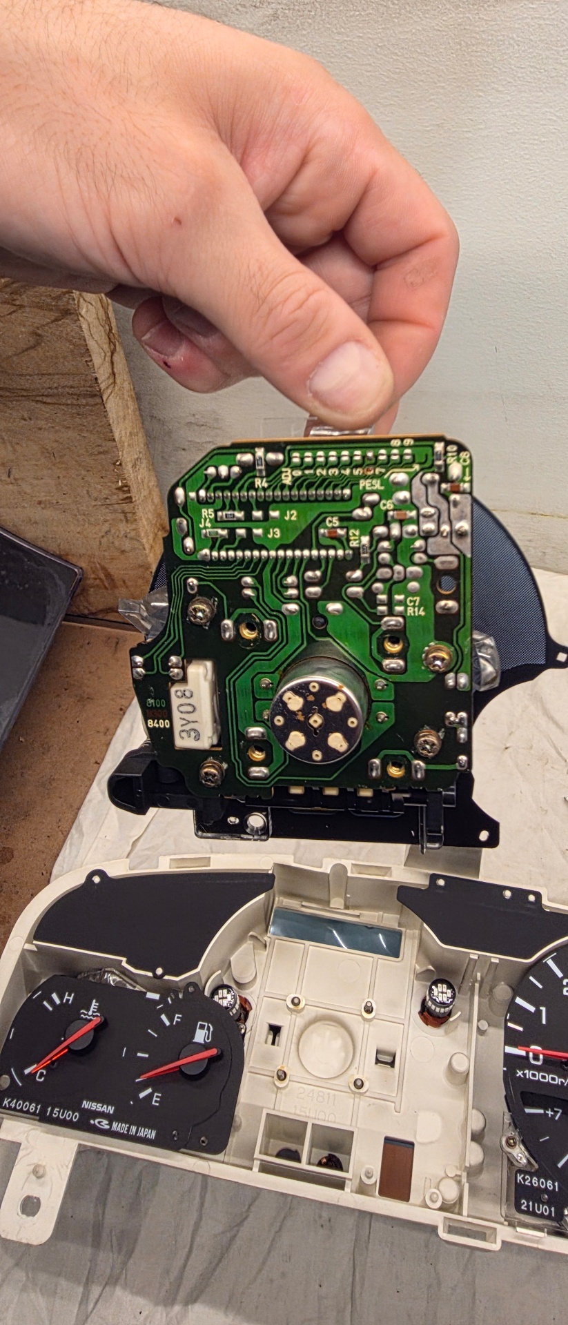 Hand holding exposed Nissan cluster PCB showing solder joints and traces above the cluster housing